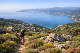 Hiker on a narrow trail through maquis with scattered granite rocks, overlooking the Gulf of Ajaccio under soft morning light