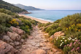 Rocky coastal footpath through native maquis leading to a quiet, wild beach at Golfe de Lava in soft morning light, with blue sea and sky in the distance.