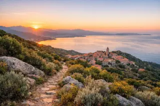 Maquis hillside path above Villanova at sunrise with panoramic view over Ajaccio gulf, wild scrub in foreground, village and sea in calm Mediterranean light.
